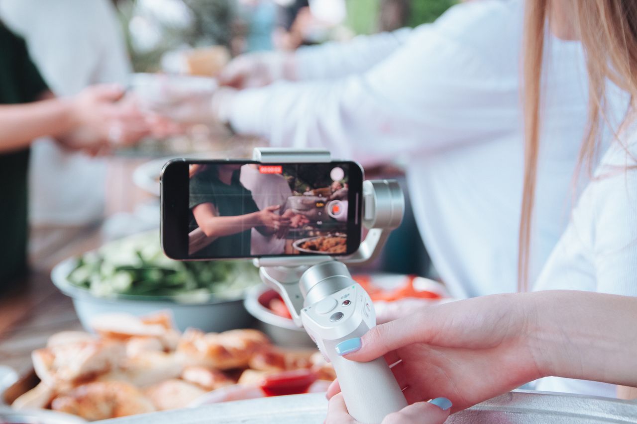 A person uses a smartphone gimbal to film a food event with various dishes.