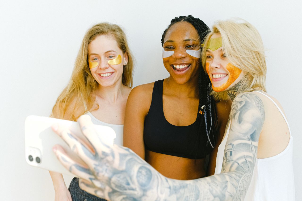 Group of happy women laughing while having fun with cosmetic eyes patches and mask for face while standing together and taking selfie on smartphone